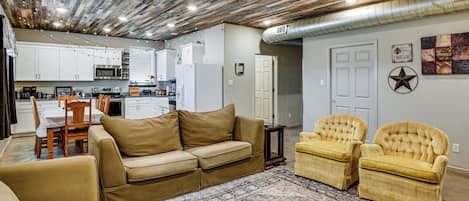 A living room with a beige sofa, two armchairs, a patterned rug, and a kitchen in the background.