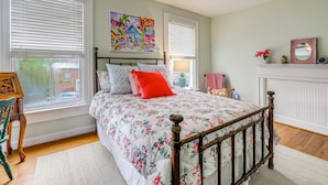 A bedroom with a bed, a wooden headboard, a window with blinds, a painting on the wall, and a fireplace mantel with a mirror and flowers. - 4BR Historic Home Located in Downtown New Bern (New Bern)