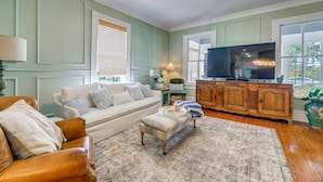 A living room with a sofa, ottoman, wooden cabinet, and a television. - 4BR Historic Home Located in Downtown New Bern (New Bern)