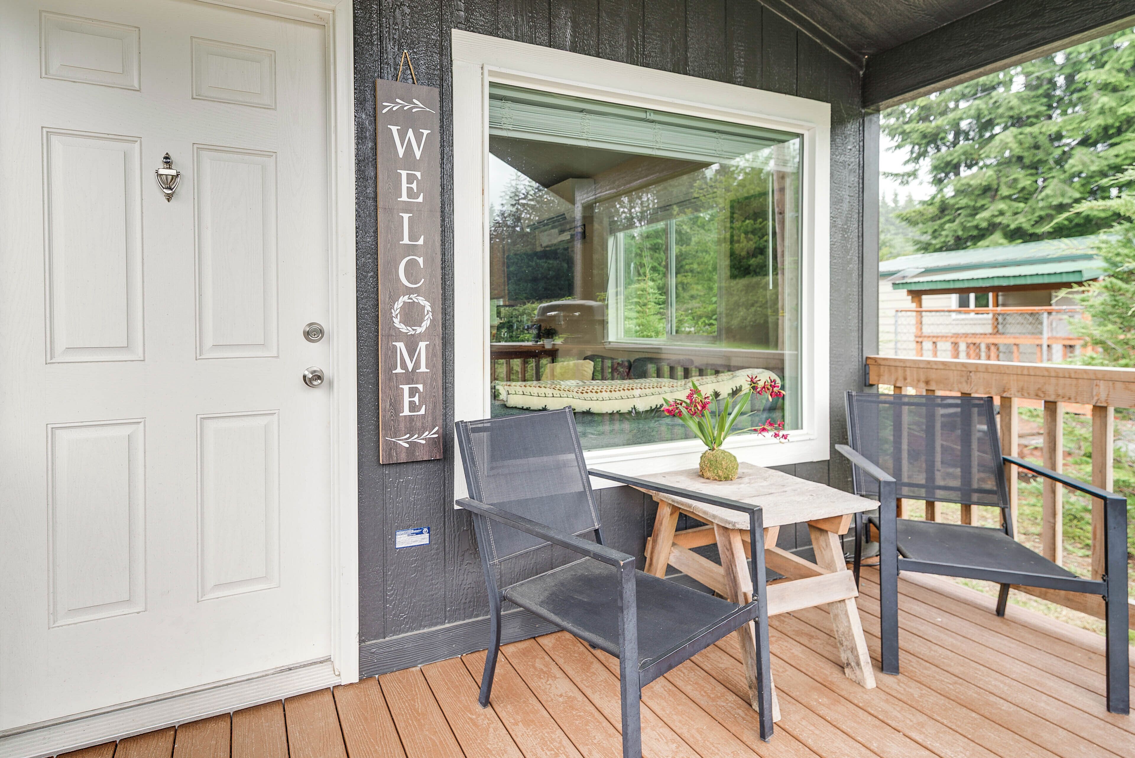 A wooden deck with a table and chairs, a white door with a 'WELCOME' sign, and a window overlooking greenery.