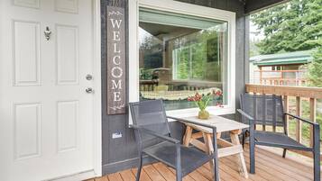 A wooden deck with a table and chairs, a white door with a 'WELCOME' sign, and a window overlooking greenery.