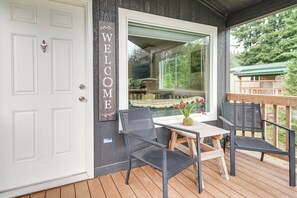 A wooden deck with a table and chairs, a white door with a 'WELCOME' sign, and a window overlooking greenery.