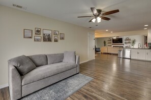 Ein geräumiges Wohnzimmer mit grauem Sofa, Holzboden, Deckenventilator und gerahmten Bildern an der Wand.