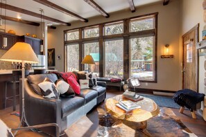 A modern living room with a leather sofa, a coffee table, and large windows offering a view of the outdoors. - Great for Families! Woodsy Sanctuary in Mc Leod (McLeod)