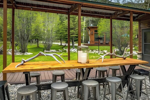 Une terrasse extérieure en bois, avec une longue table et de nombreux tabourets, entourée d’un paysage naturel composé d’arbres et d’un petit bâtiment en arrière-plan.