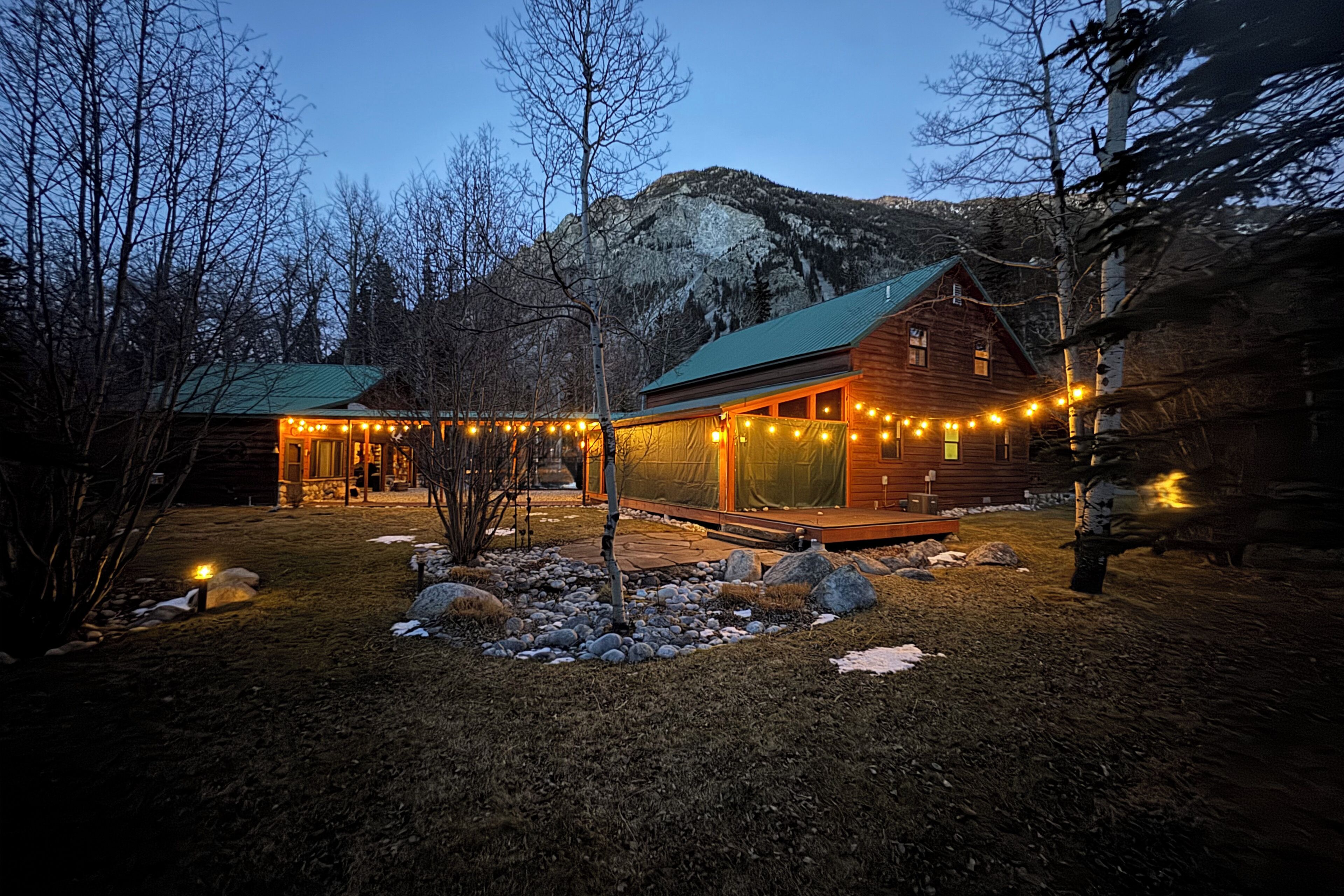 Une cabane en rondins surmontée d’un toit végétalisé, entourée d’un sentier en pierre et illuminée de guirlandes lumineuses.