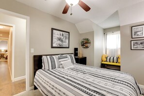 Ein Schlafzimmer mit Bett, Deckenventilator, gerahmtem Bild an der Wand und kleinem Tisch mit gelbem Kissen.