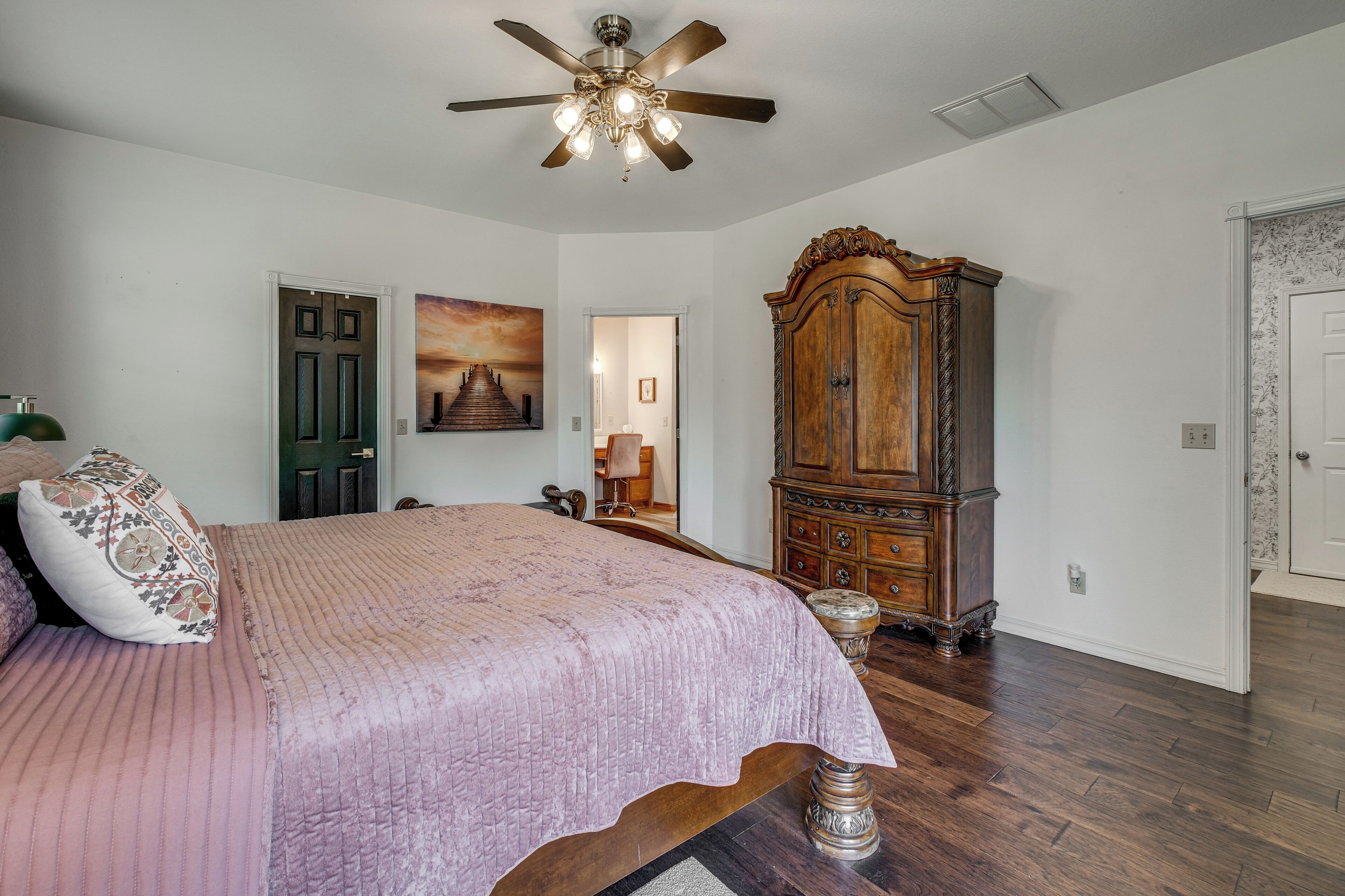 A bedroom with a bed, a ceiling fan, a wooden wardrobe, and a painting on the wall.