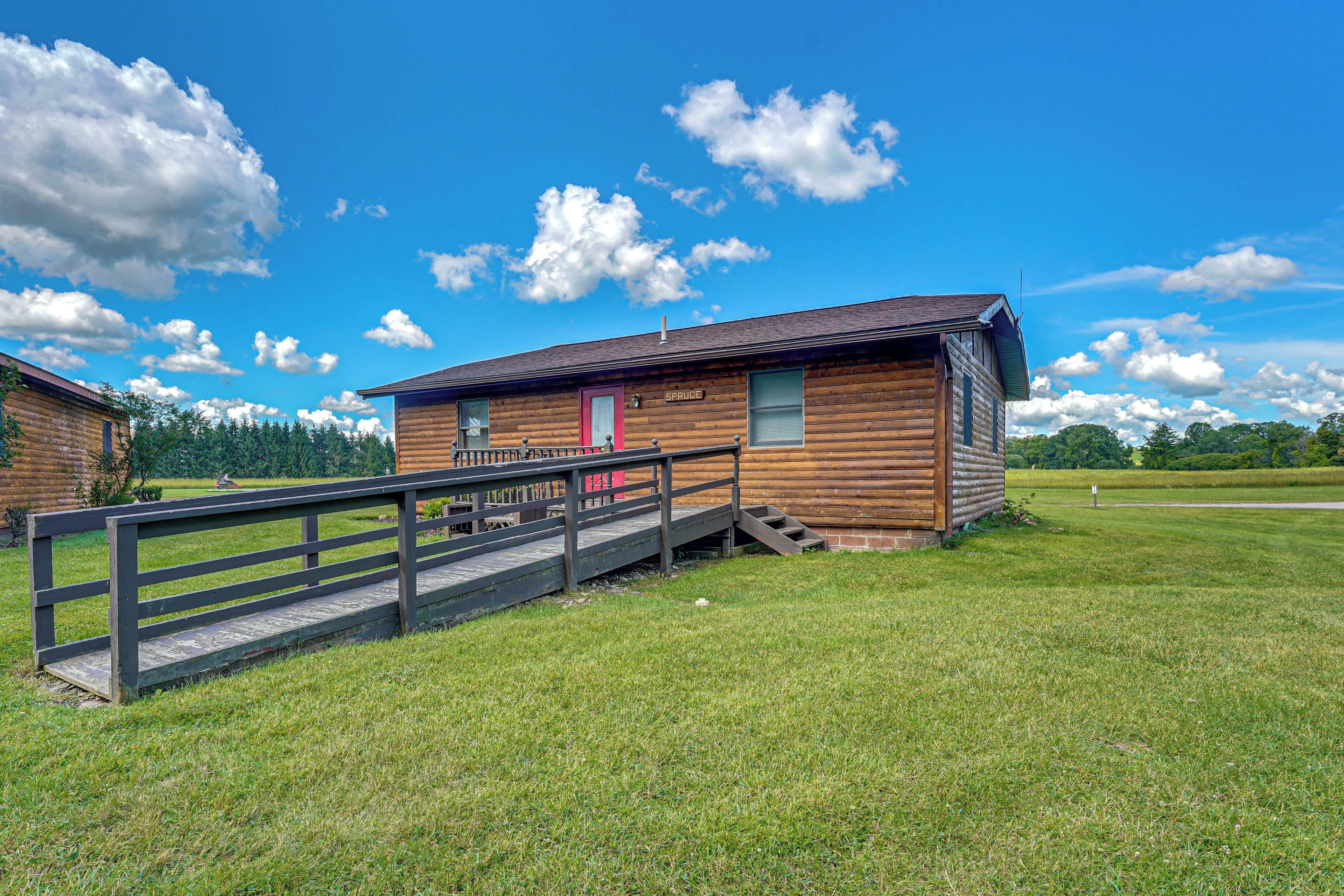 A wooden cabin with a red door, a wooden deck, and a clear blue sky with clouds.