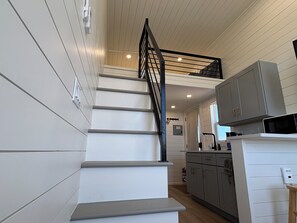 A modern kitchen with white cabinetry, a black staircase, and a small dining area. - Oceanfront Tiny Retreat On Wheels, Fall Package + 2 Bikes (Holden Beach)
