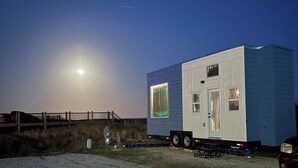 A mobile home with a large window and a door, parked on a gravel surface with a fence and a bridge in the background. - Oceanfront Tiny Retreat On Wheels, Fall Package + 2 Bikes (Holden Beach)