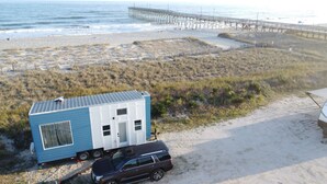 A small blue trailer with a white section, parked on a sandy beach with a beach and pier in the background. - Oceanfront Tiny Retreat On Wheels, Fall Package + 2 Bikes (Holden Beach)
