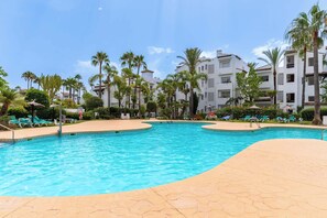 A swimming pool with lounge chairs and palm trees, surrounded by multi-story buildings. - Costalita 2BR, Pool, Garage & Direct Beach Access (Estepona)