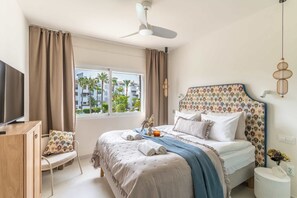 Ein Schlafzimmer mit Bett, Vorhängen, Deckenventilator, Fernseher, Sessel und einem Fenster mit Blick auf Palmen und Gebäude.