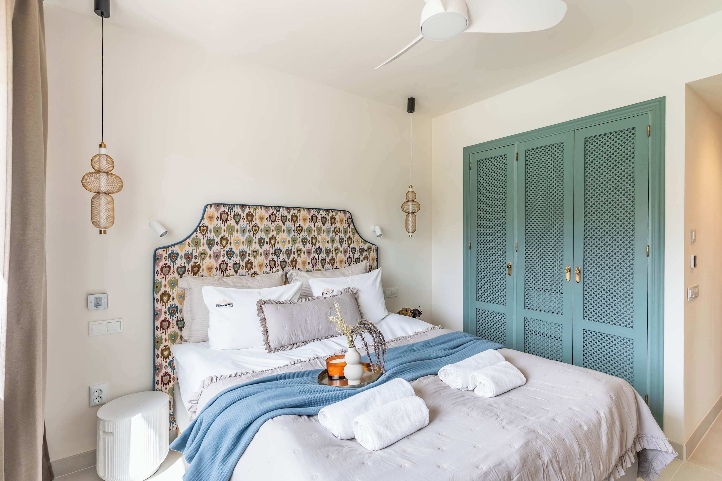 Un dormitorio con un cabecero estampado, una cama con ropa blanca y una manta azul, junto con un juego de toallas.