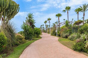 A paved pathway flanked by various plants and palm trees, leading towards a building with a tiled roof. - Costalita 2BR, Pool, Garage & Direct Beach Access (Estepona)