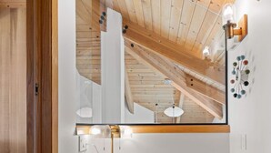 Quarto com teto de madeira, toucador branco com espelho, cadeira verde e relógio fixado na parede.