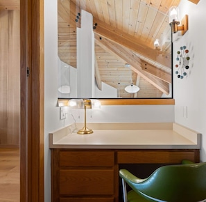 A room with a wooden ceiling, a white vanity with a mirror, a green chair, and a wall-mounted clock. - Unit 35C Lot 8 Oceanhaus (The Sea Ranch)