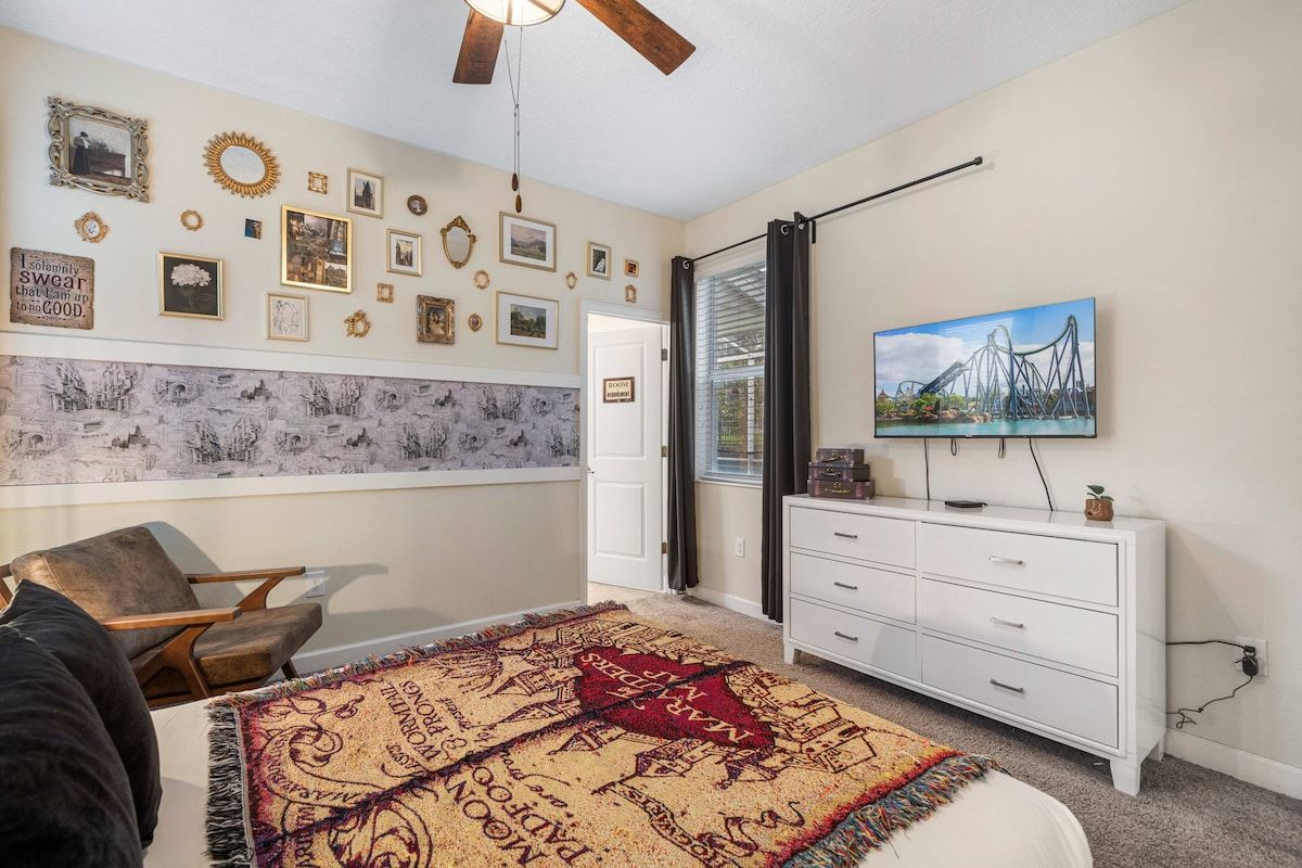 A bedroom with a bed, a white dresser, a television, and a wall decorated with various framed pictures and a mirror.