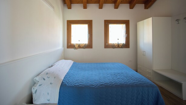 A bedroom with a bed, two windows, a wardrobe, and wooden beams on the ceiling.