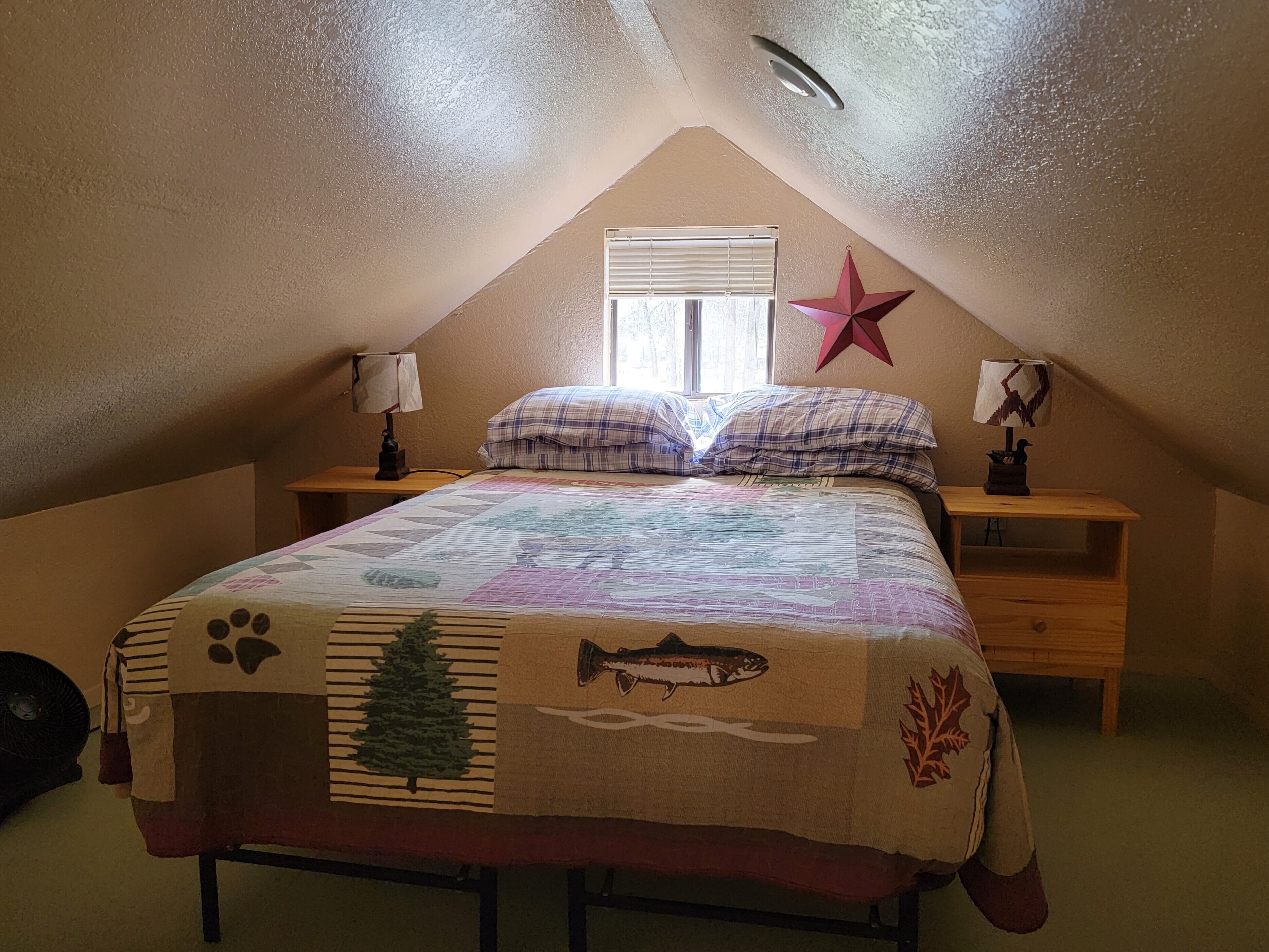 A bedroom with a sloped ceiling, a bed with a quilt, two bedside tables with lamps, a window with blinds, and a star decoration on the wall.