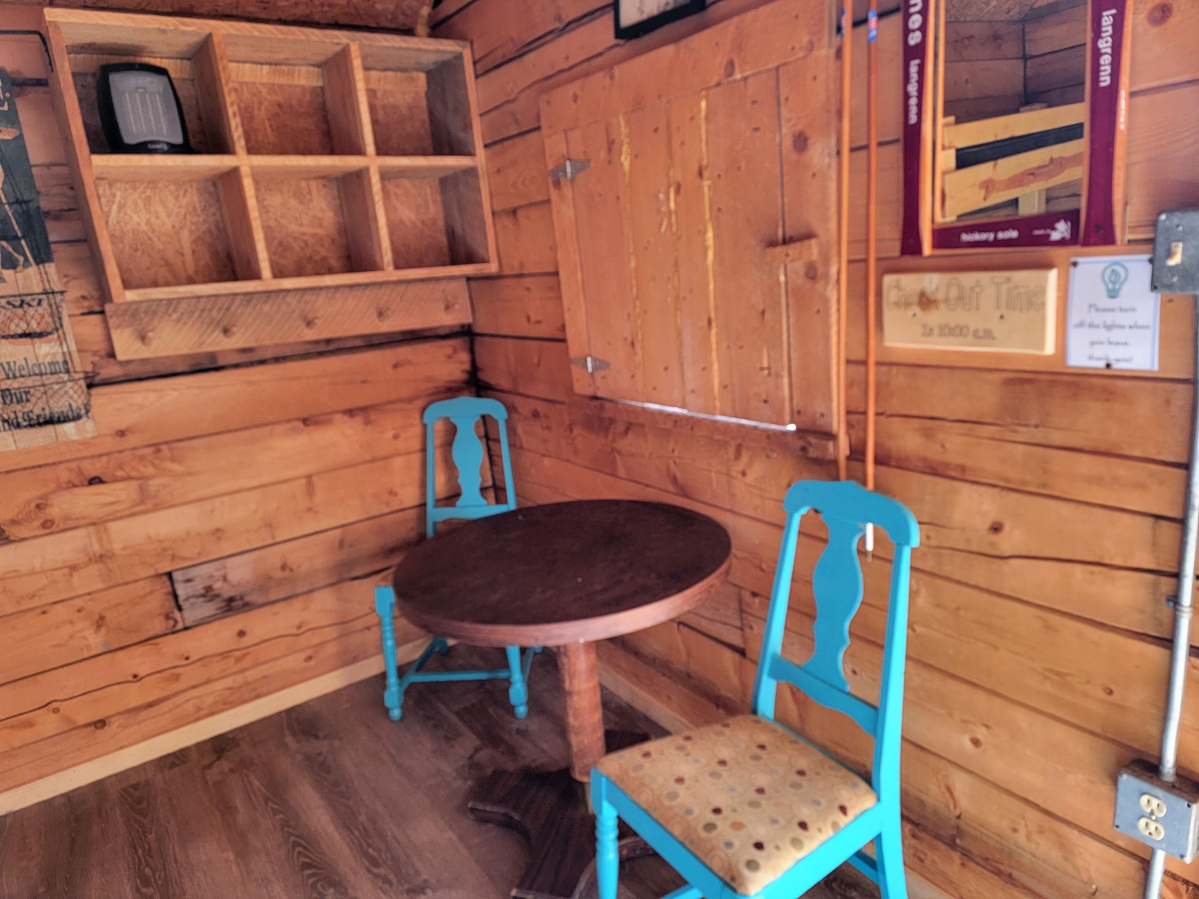 A wooden interior with a round table and two chairs, a shelf with items, and a wall-mounted sign.