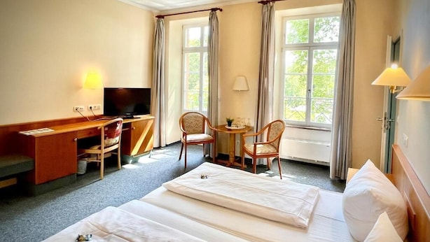 Habitación de hotel con dos camas, un escritorio con televisor, una silla, una mesita y ventanas grandes con cortinas.