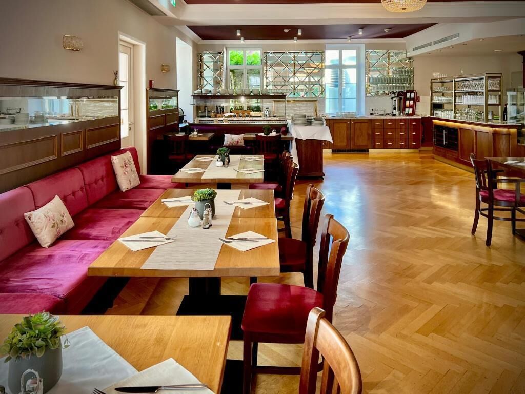 A spacious dining area with a long wooden table, red upholstered seating, and a kitchen in the background.