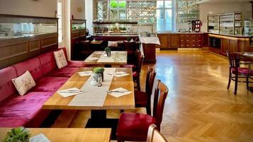 Un vaste espace dédié à la salle à manger, avec une longue table en bois, des sièges rembourrés de couleur rouge et une cuisine en arrière-plan.