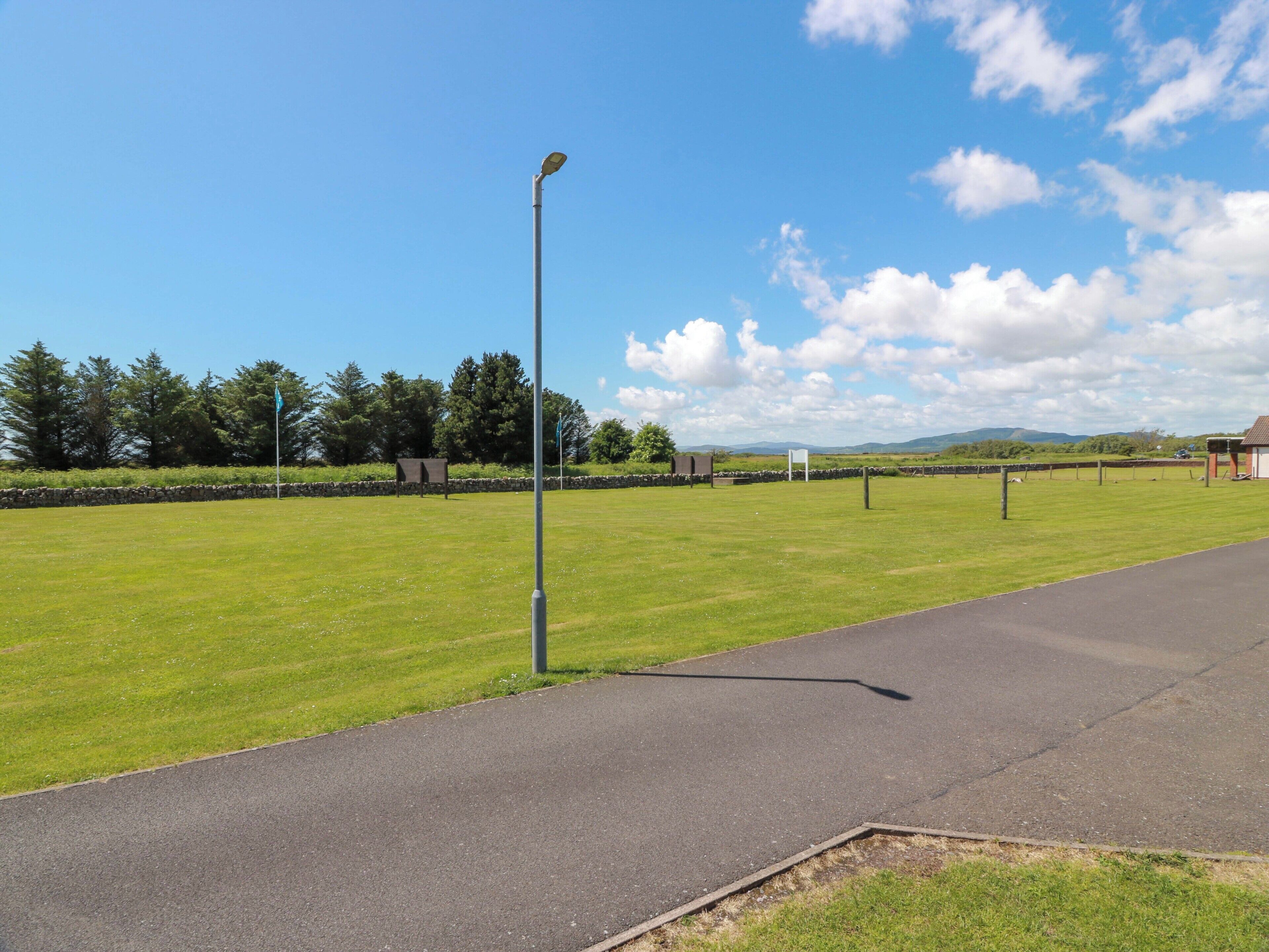 A grassy field with a paved path, a street lamp, and a stone wall in the background.