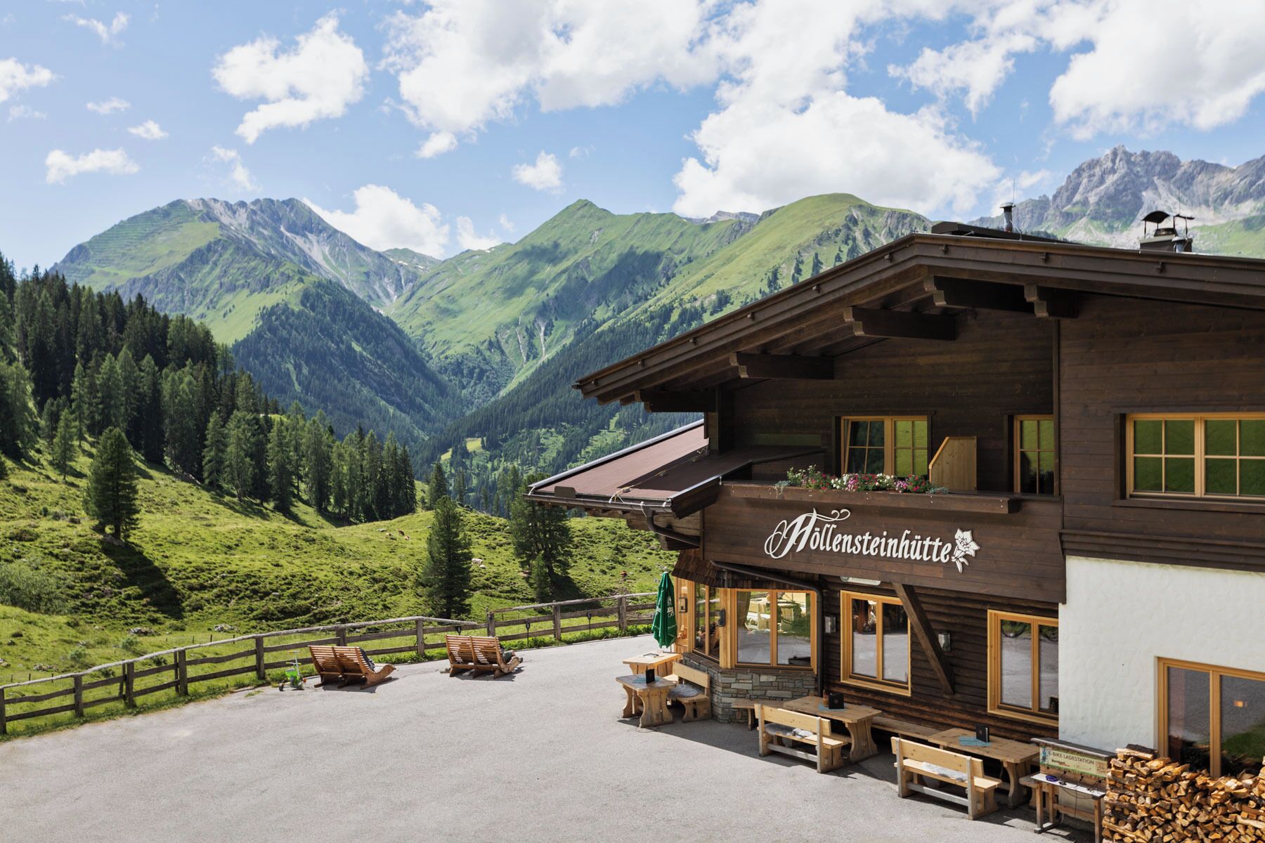 A wooden building with a sign reading 'Höllsteinhütte' in a mountainous area with outdoor seating and a view of the mountains.