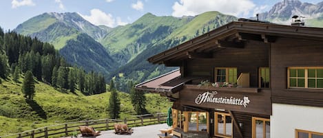 A wooden building with a sign reading 'Höllsteinhütte' in a mountainous area with outdoor seating and a view of the mountains.