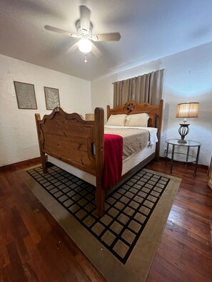 Ein Schlafzimmer mit einem hölzernen Bett, einem Deckenventilator, einem kleinen Tisch mit Lampe und einem Fenster mit Vorhängen.