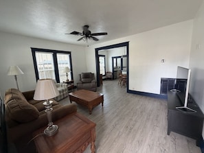 A living room with a sofa, coffee table, and lamp. There is a ceiling fan, a fireplace, and a television. - Built for Work Crews & Extended Business Stays. (Gainesville)