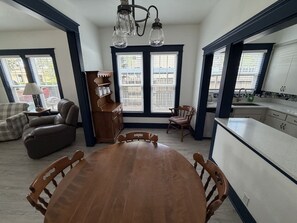 A dining area with a wooden table and chairs, a kitchen with white cabinets, and a window with blinds. - Built for Work Crews & Extended Business Stays. (Gainesville)