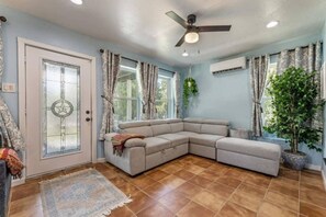 Una sala de estar con un sofá modular, un ventilador de techo, una planta en maceta y un ventanal con cortinas.