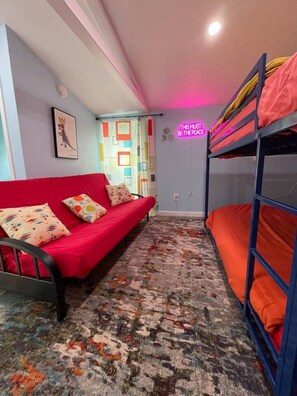 Ein Zimmer mit einem Etagenbett, einem roten Schlafsofa, einem gemusterten Teppich, einem gerahmten Bild an der Wand und einer Neonreklame.