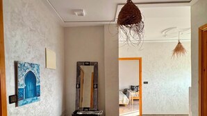 Un couloir avec un sol carrelé, une porte en bois, un miroir et un luminaire suspendu.