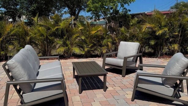 Een terras buiten met drie grijze loungestoelen en een kleine tafel, omgeven door diverse planten en bomen.