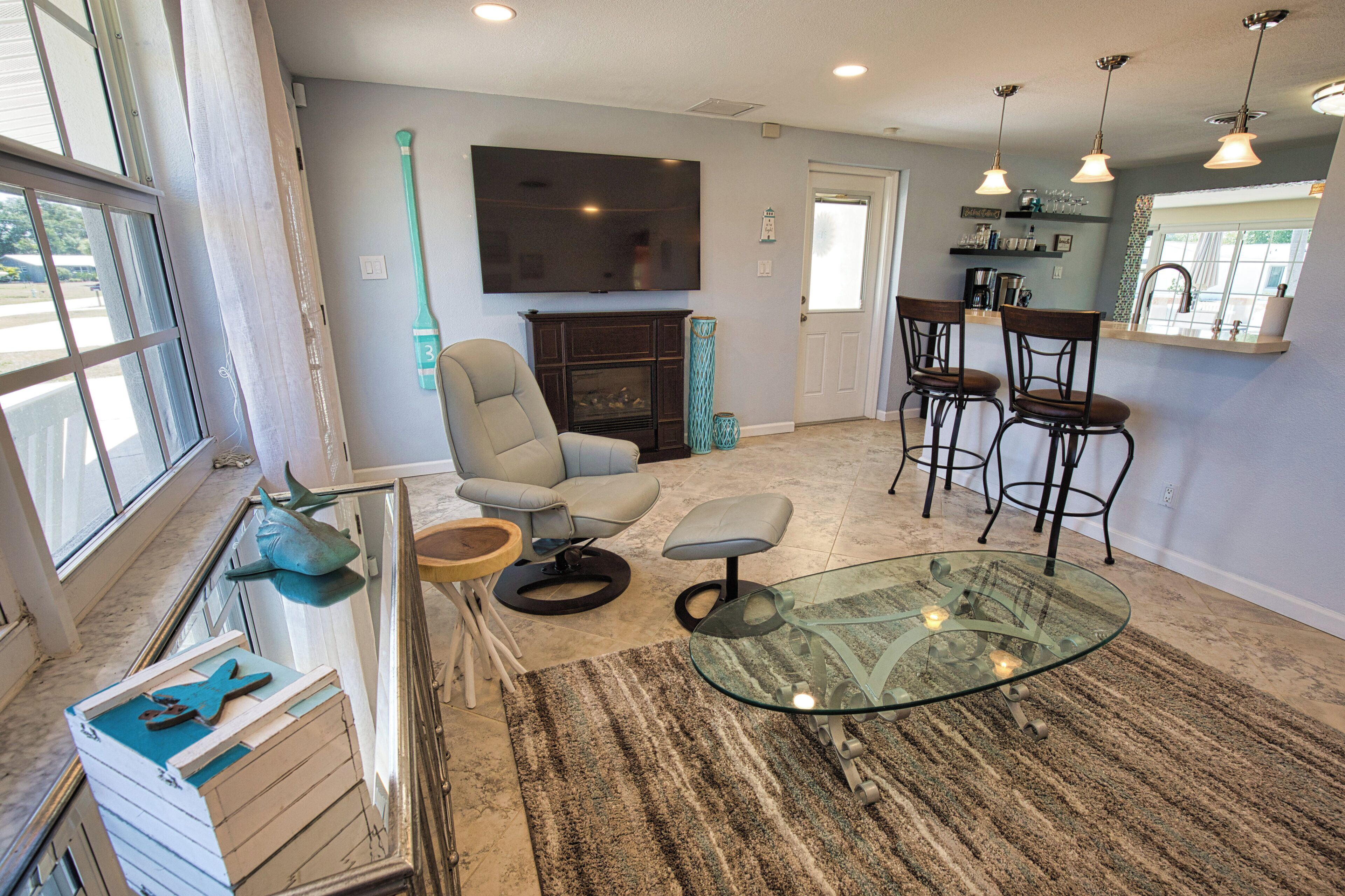 A modern living room with a glass coffee table, a comfortable armchair, a fireplace, and a kitchen area with bar stools.
