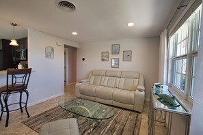 Ein Wohnzimmer mit Glastisch, einem beigefarbenen Sofa, einem Essbereich mit Stuhl und einer Küchenzeile mit Schränken und einer Mikrowelle.