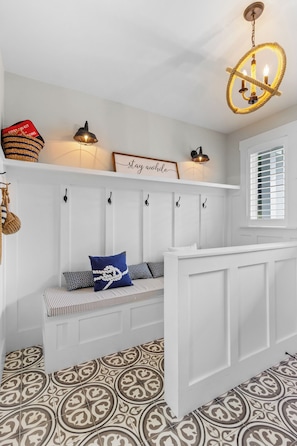 A white interior with a bench, wall-mounted hooks, a chandelier, and patterned floor tiles.
