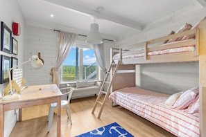 A room with bunk beds, a desk with a lamp, a window with curtains, and a view of greenery.