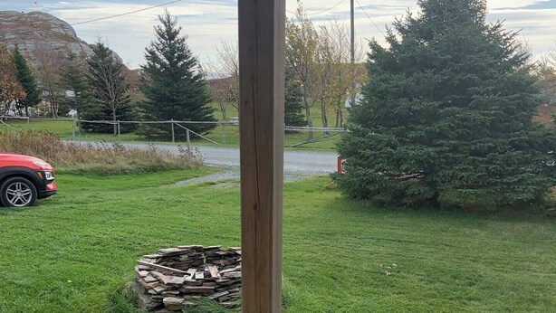 A wooden deck with a view of a grassy yard, a fire pit, and a red vehicle in the distance. - Escape to The Blue Bird – a cozy 2-bedroom cottage with hot tub and partial ocean views! Just 5 mins from Carbonear & 15 mins to Salmon Cove Sands. (Flat Rocks)
