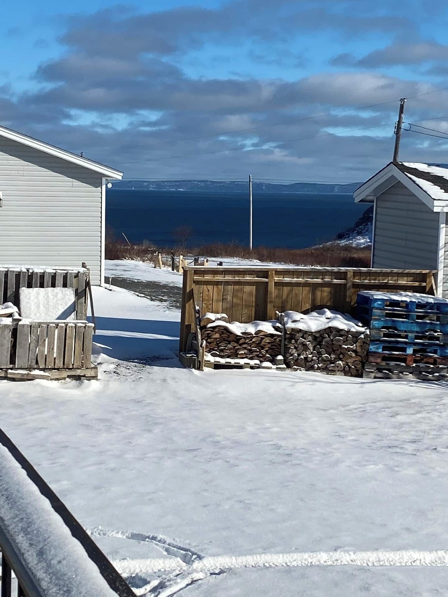 A snowy outdoor area with a wooden fence, a stack of firewood, and a small shed.