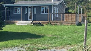 A single-story house with a front porch, a chimney, and a deck. - Escape to The Blue Bird – a cozy 2-bedroom cottage with hot tub and partial ocean views! Just 5 mins from Carbonear & 15 mins to Salmon Cove Sands. (Flat Rocks)
