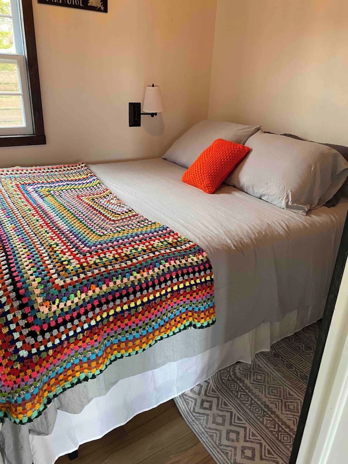 A bed with a multicolored crocheted blanket, a single orange pillow, and a wall-mounted lamp.