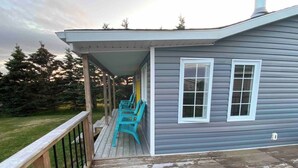 A house with a covered porch, a wooden deck, and two windows. - Escape to The Blue Bird – a cozy 2-bedroom cottage with hot tub and partial ocean views! Just 5 mins from Carbonear & 15 mins to Salmon Cove Sands. (Flat Rocks)