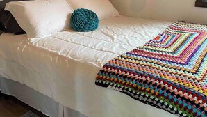 A bed with a white duvet, a teal knitted pillow, and a multicolored crocheted blanket. - Escape to The Blue Bird – a cozy 2-bedroom cottage with hot tub and partial ocean views! Just 5 mins from Carbonear & 15 mins to Salmon Cove Sands. (Flat Rocks)