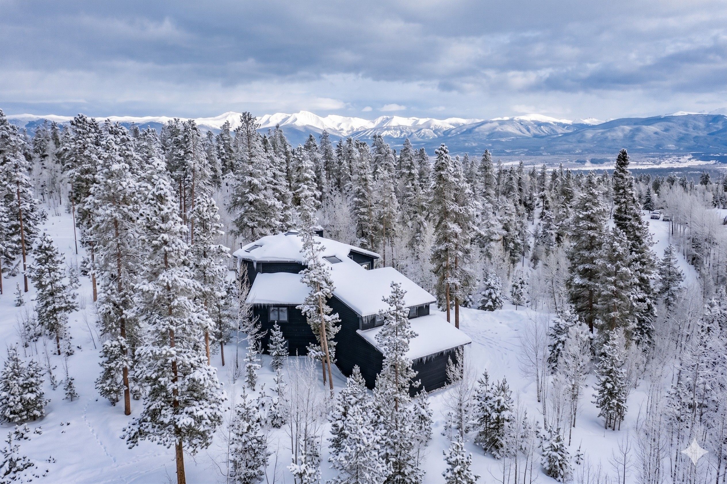 A cabin surrounded by a dense forest of snow-covered trees, with a mountain range in the background.